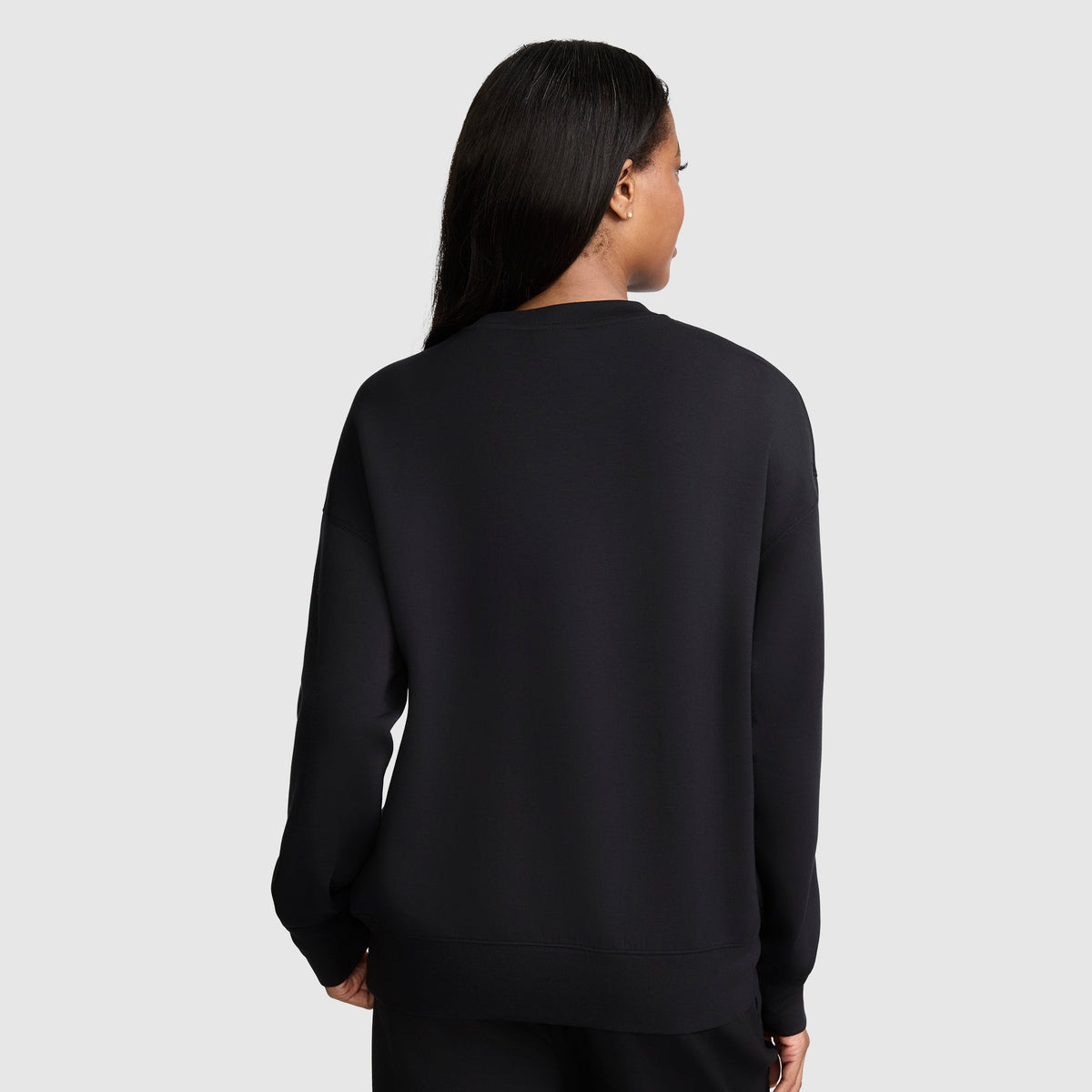A woman with long dark hair stands with her back to the camera against a light gray background, wearing black pants and a Champion Crewneck Pullover Sweatshirt. The plain black modal scuba design of the sweatshirt creates an athletic silhouette.
