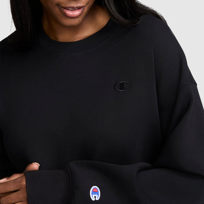 A person is shown from the shoulders to mid-torso wearing the Champion Crewneck Pullover Sweatshirt in black, featuring an embroidered C logo on the chest and a second logo on the sleeve, showcasing a classic athletic silhouette.