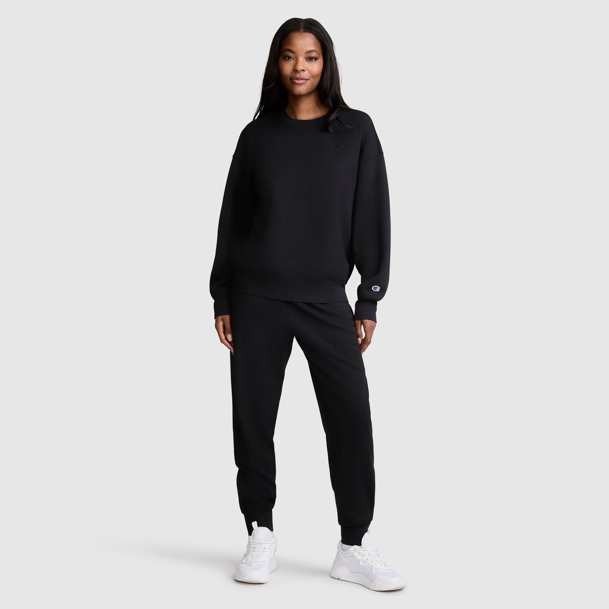 A woman stands facing forward, wearing a Champion Crewneck Pullover Sweatshirt in black with matching joggers and white sneakers, showcasing an athletic silhouette against a plain light gray background.