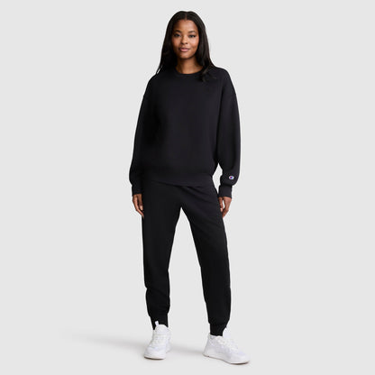A woman stands facing forward, wearing a Champion Crewneck Pullover Sweatshirt in black with matching joggers and white sneakers, showcasing an athletic silhouette against a plain light gray background.