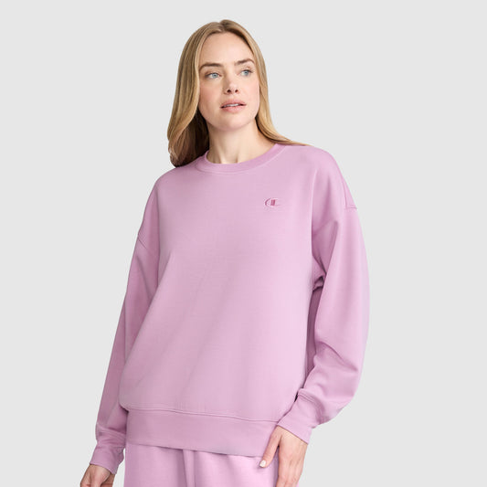 A woman stands against a plain light gray background, looking slightly to the side with a neutral expression, dressed in a loose-fitting pastel pink Champion Crewneck Pullover Sweatshirt and matching pants. The athletic silhouette of her Champion top adds a modern touch to her laid-back look.