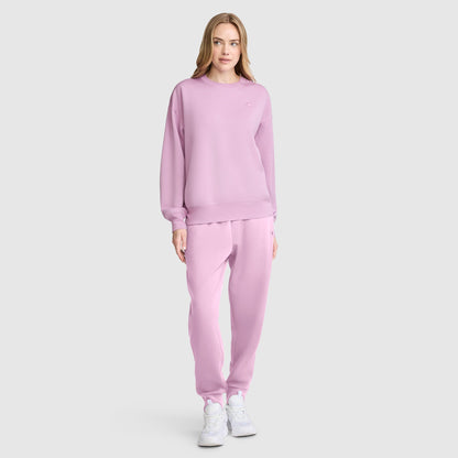 A woman stands facing forward against a plain light gray background, wearing a matching light pink Champion Crewneck Pullover Sweatshirt and sweatpants with white sneakers.