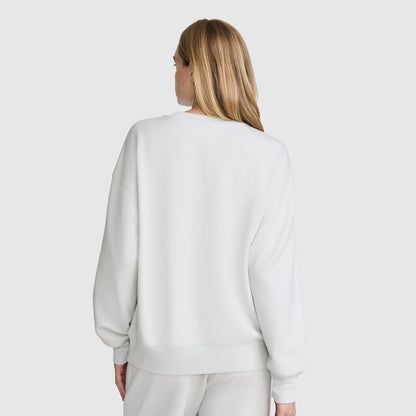 A woman with long, light brown hair is seen from behind against a plain, light background, wearing a loose-fitting Champion Crewneck Pullover Sweatshirt paired with matching white pants.