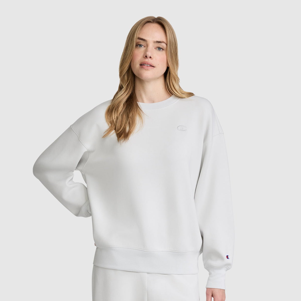 A woman with long blonde hair wears a loose-fitting white Champion Crewneck Pullover Sweatshirt and matching white pants, standing against a plain light gray background with one hand on her hip.