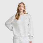 A woman with long blonde hair wears a loose-fitting white Champion Crewneck Pullover Sweatshirt and matching white pants, standing against a plain light gray background with one hand on her hip.