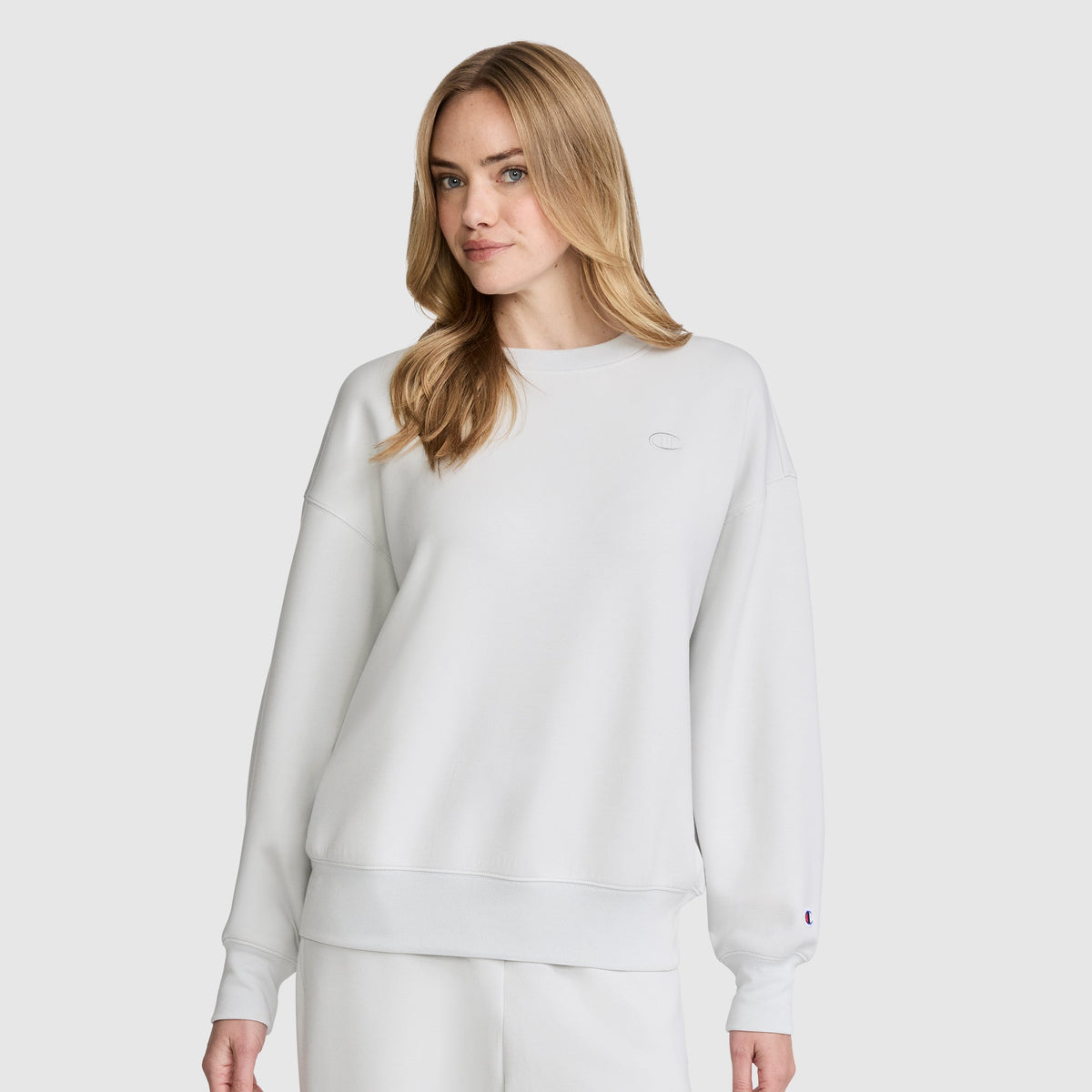 A woman with long blonde hair wears a plain white Champion Crewneck Pullover Sweatshirt and matching white pants, standing against a light gray background.
