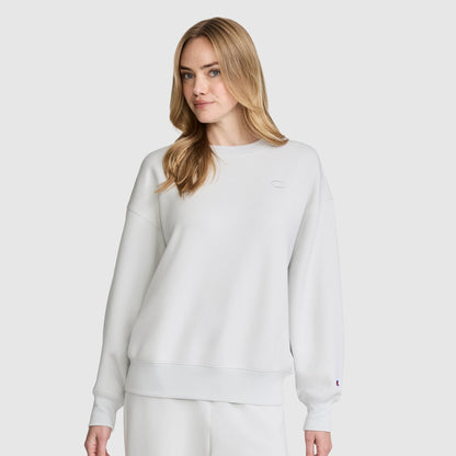A woman with long blonde hair wears a plain white Champion Crewneck Pullover Sweatshirt and matching white pants, standing against a light gray background.