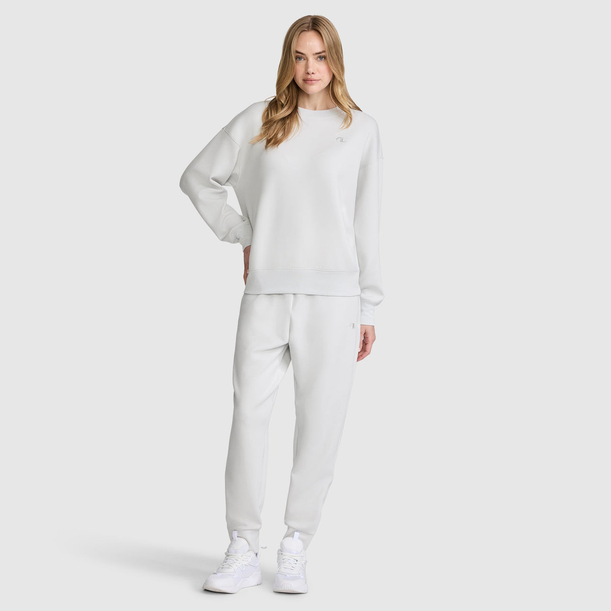 A woman with long, blonde hair stands against a plain background, wearing a matching light gray Champion Crewneck Pullover Sweatshirt and sweatpants set with white sneakers. She looks relaxed and casual.