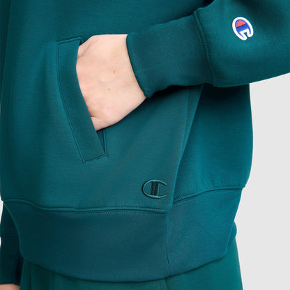 A person wearing the Champion Quarter-Zip Pullover in teal with one hand in the front pocket. The sporty style of this sweater is highlighted by the Champion logo patch on the sleeve and embroidered branding near the pocket.