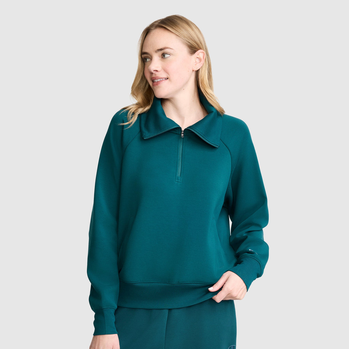 Wearing the Champion Quarter-Zip Pullover in teal stretch poly-modal, paired with matching pants, a person stands against a plain light gray background, looking to the side and smiling softly.