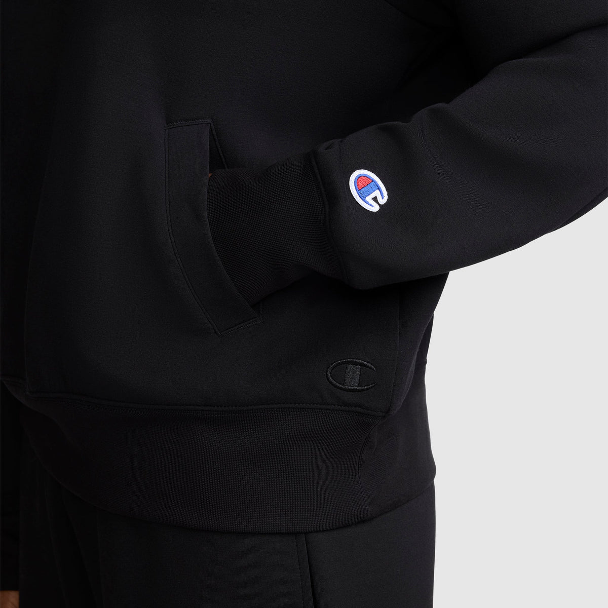 A person is wearing a Champion Quarter-Zip Pullover in black, with one hand tucked into the front pocket and the Champion logo patch visible on the sleeve. The photo is cropped to show the athletic pullover from shoulder to upper thigh.