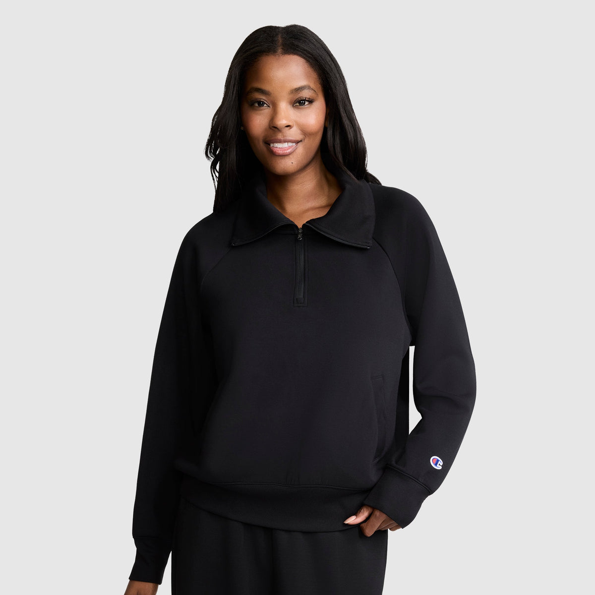 A woman stands against a plain light gray background, smiling with one hand relaxed by her side, wearing a black Champion Quarter-Zip Pullover and matching pants.