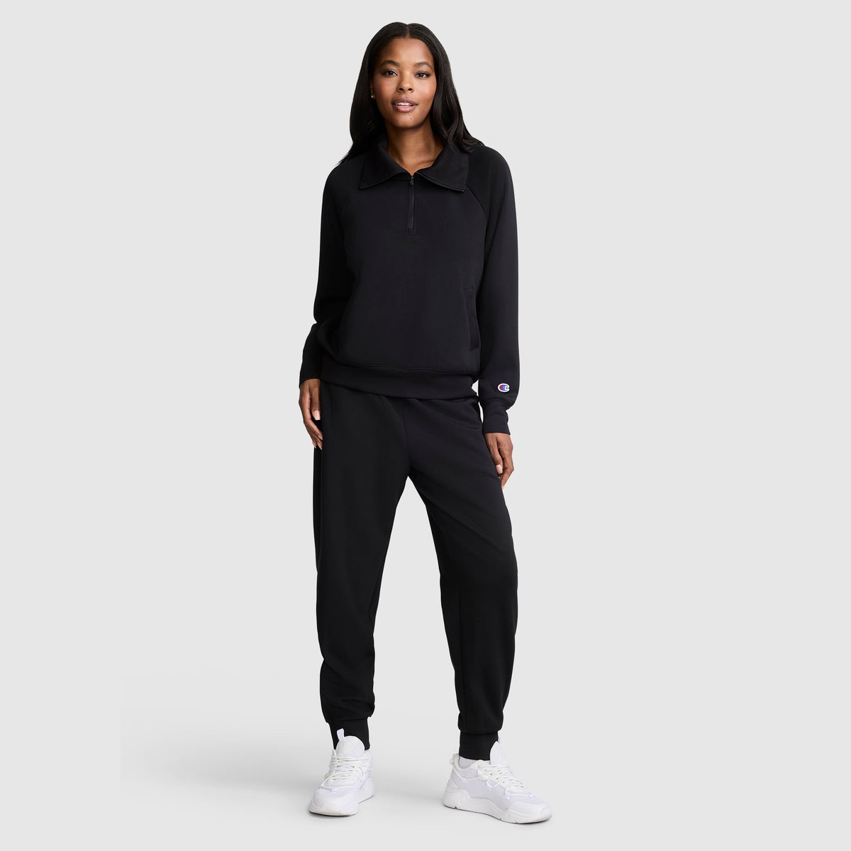 A woman stands against a plain light gray background, dressed in a sleek black Champion Quarter-Zip Pullover paired with matching black joggers and white sneakers. The outfit highlights the classic style and comfortable, sporty elegance of the Champion Quarter-Zip Pullover.
