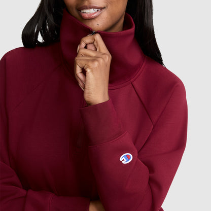 A woman, smiling gently, zips up the high collar of a maroon Champion Quarter-Zip Pullover. Only her lower face and upper body are visible against a plain background.