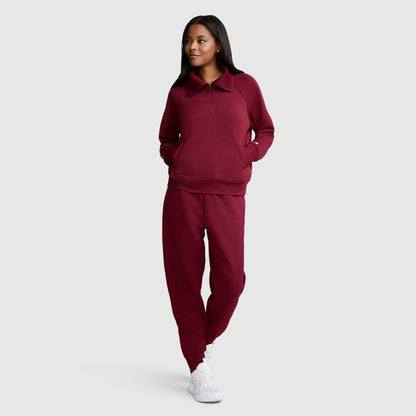 A woman wearing a Champion Quarter-Zip Pullover in maroon stretch scuba blend with matching pants and white sneakers stands with her hands in her pockets, smiling and looking to the side against a plain light gray background.