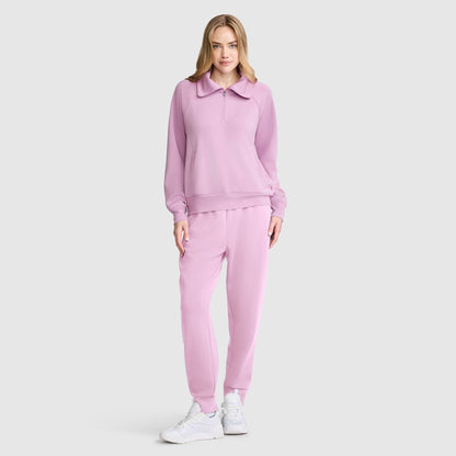 A woman stands facing forward against a plain light gray background, wearing a matching light pink Champion Quarter-Zip Pullover tracksuit and white sneakers. She is smiling softly, with her left hand in her pocket.