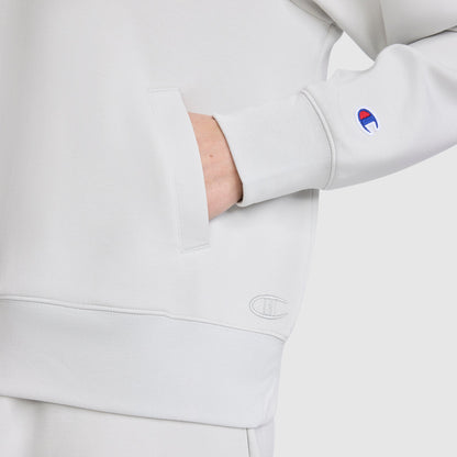 A person is wearing a light gray Champion Quarter-Zip Pullover with one hand in the front pocket. The Champion logo appears on the sleeve and near the waistband. This athletic pullover by Champion is crafted from soft, stretch poly-modal fabric for comfort and flexibility. The background is plain white.