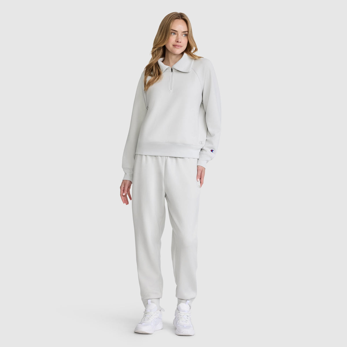 A woman stands against a plain background, wearing the Champion Quarter-Zip Pullover in light gray with a collar, paired with matching jogger pants made from soft, stretch poly-modal fabric. She has long, wavy hair and completes the look with white sneakers.