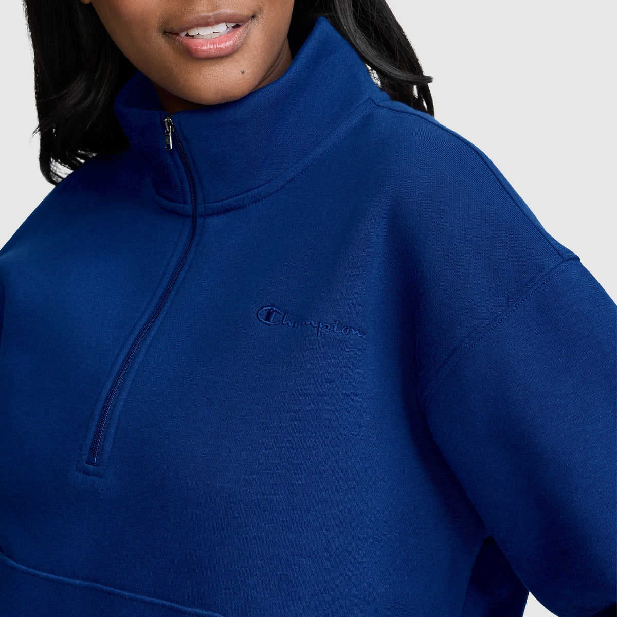 A woman wearing a royal blue Champion Fleece Mockneck Quarter-Zip, featuring the iconic Champion logo on the chest, stands against a plain light background and smiles softly, embodying timeless sport style.