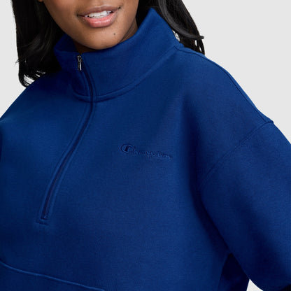 A woman wearing a royal blue Champion Fleece Mockneck Quarter-Zip, featuring the iconic Champion logo on the chest, stands against a plain light background and smiles softly, embodying timeless sport style.
