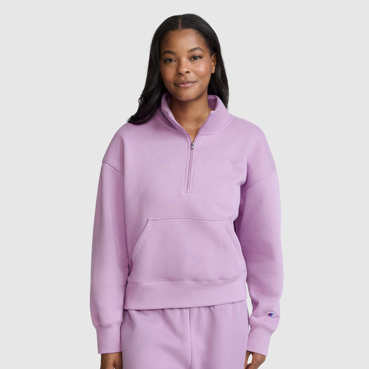 A woman stands facing forward, smiling slightly, against a plain light gray background, dressed in a light purple Champion Fleece Mockneck Quarter-Zip and matching pants that showcase a casual sport style.