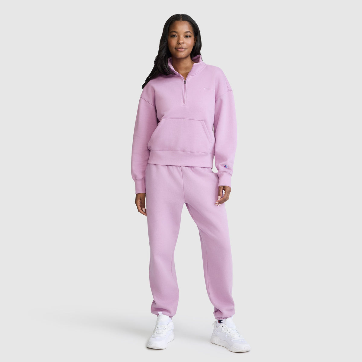 A woman stands smiling, wearing a light purple Champion Fleece Mockneck Quarter-Zip sweatshirt and matching sweatpants with white sneakers, set against a plain light gray background.