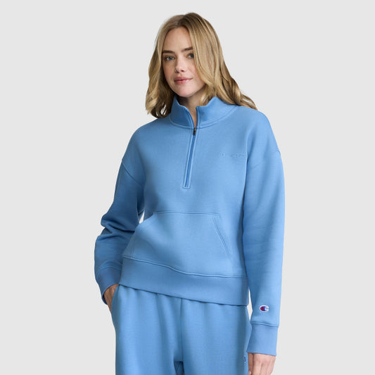 A blonde-haired woman models the Champion Fleece Mockneck Quarter-Zip in light blue with coordinating sweatpants, standing with one hand in her pocket against a plain light background for a relaxed sporty look.