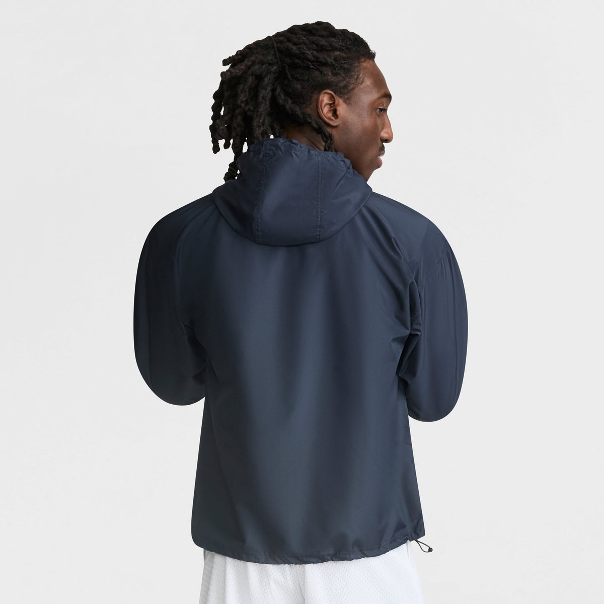 A person with dark, shoulder-length twists stands with their back to the camera against a plain light background, wearing the Champion Stadium Packable Jacket, Script Logo in dark navy and white shorts.