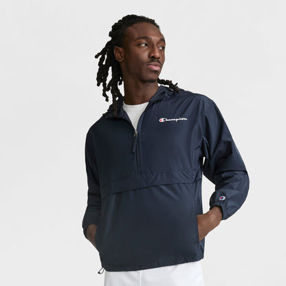 A man with long braided hair wears a navy Champion Stadium Packable Jacket with Script Logo and half-zip, hands in pockets. He looks left while standing against a plain white background.