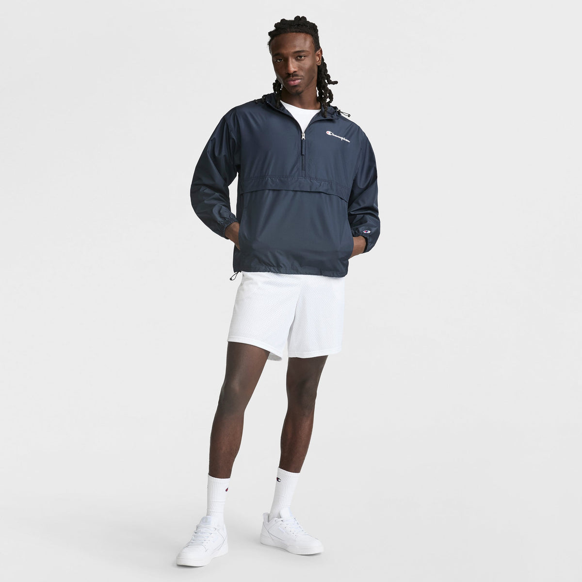 A man stands against a plain white background wearing the Champion Stadium Packable Jacket with Script Logo in navy blue, paired with white athletic shorts, socks, and sneakers. His hands are relaxed by his sides.