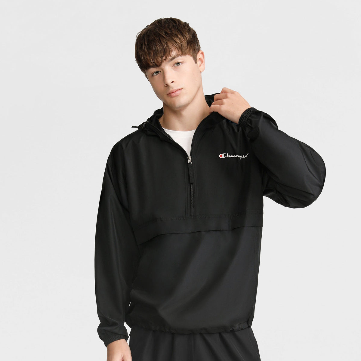 A young man with short brown hair wears a black Champion Stadium Packable Jacket featuring a script logo on the chest and front zipper, posing with one hand on his shoulder against a plain light background.