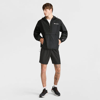 A young man in a black Champion Stadium Packable Jacket with Script Logo, black shorts, white socks, and black sneakers stands against a white background. He has visible leg tattoos and looks to the side with hands in his jacket pockets.