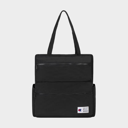 The Champion Explore Tote is crafted from durable crinkle nylon in black and features two zippered front pockets, convenient side pockets, and a small Champion logo patch on the lower right corner. Inside, a padded laptop sleeve adds functionality. The product is displayed against a plain white background.