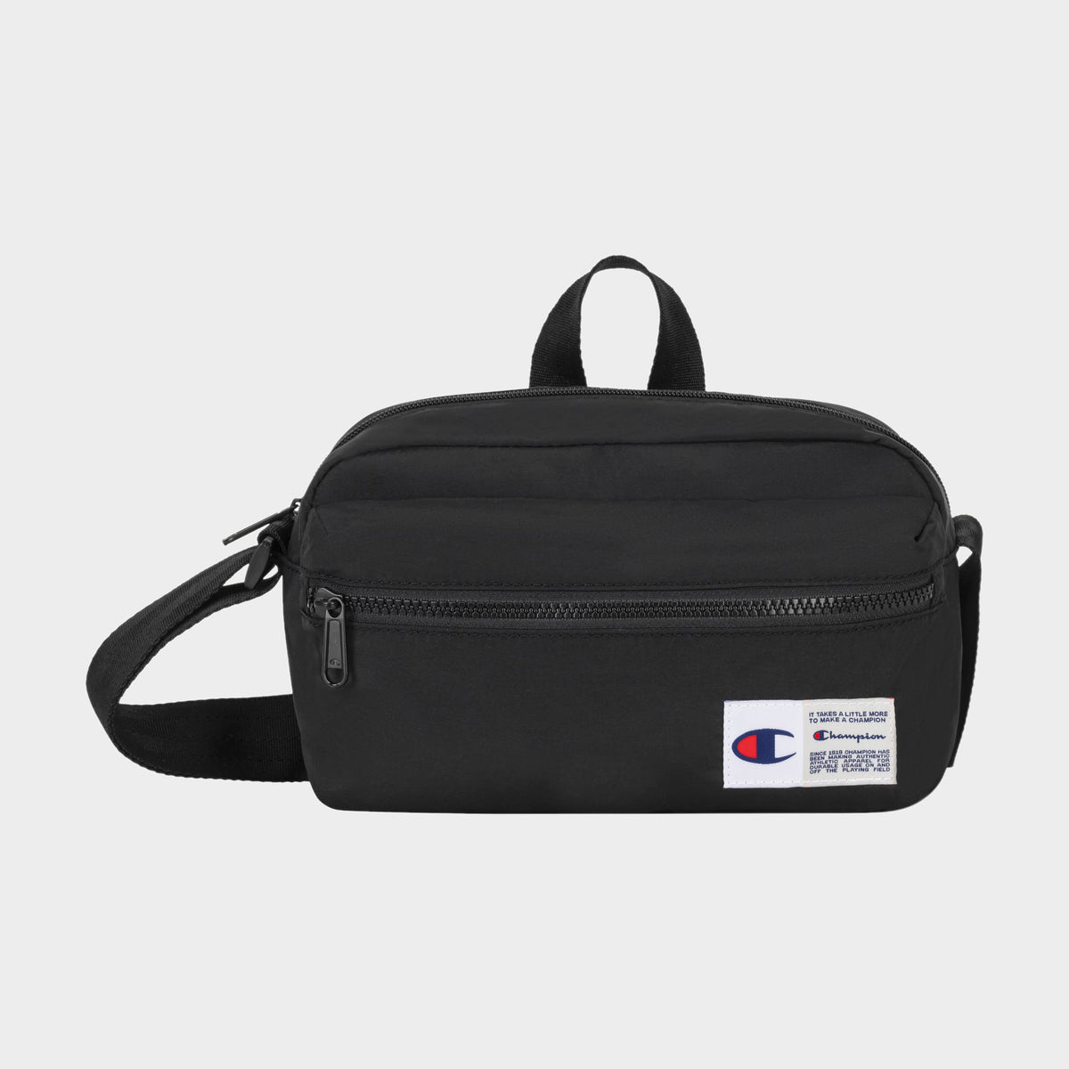 The Explore Crossbody by Champion is a black nylon bag with an adjustable strap, a front zipper pocket, and a small white Champion logo patch on the lower right corner. It is shown against a plain light background.