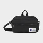 The Explore Crossbody by Champion is a black nylon bag with an adjustable strap, a front zipper pocket, and a small white Champion logo patch on the lower right corner. It is shown against a plain light background.