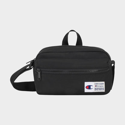 The Explore Crossbody by Champion is a black nylon bag with an adjustable strap, a front zipper pocket, and a small white Champion logo patch on the lower right corner. It is shown against a plain light background.