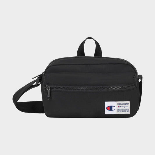 The Explore Crossbody by Champion is a black nylon bag with an adjustable strap, a front zipper pocket, and a small white Champion logo patch on the lower right corner. It is shown against a plain light background.