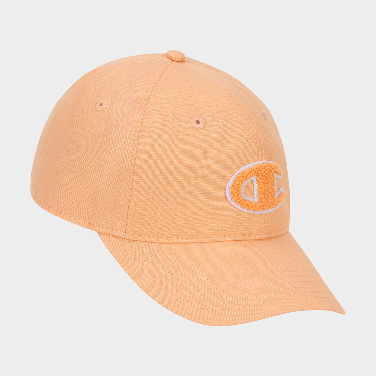 The Amber Chenille Adjustable Cap by Champion features a peach-colored, relaxed fit with a curved brim and an embroidered chenille “C” patch on the front left panel, displayed against a plain white background.