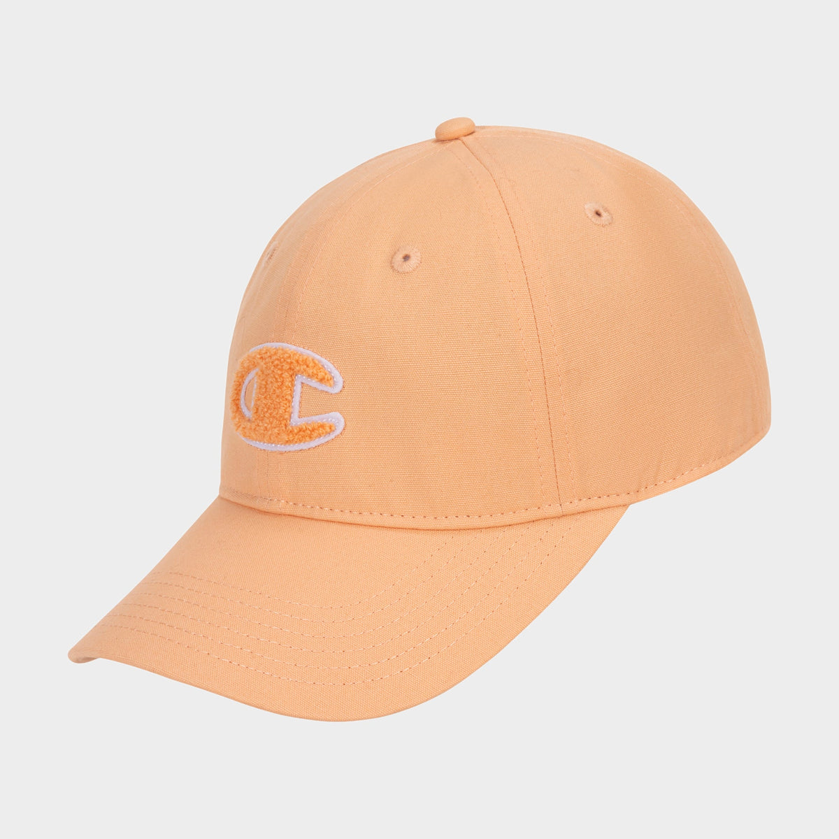 The Amber Chenille Adjustable Cap by Champion is peach-colored with a relaxed fit, featuring a curved brim and a white and yellow chenille C logo patch embroidered on the front. The cap is displayed against a plain white background.