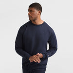 A man wears a navy Champion Powerblend Crewneck Sweatshirt, C Logo - Big & Tall, and matching pants, standing against a white background with his hands loosely clasped, looking to his right.