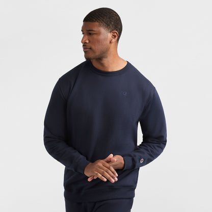 A man wears a navy Champion Powerblend Crewneck Sweatshirt, C Logo - Big & Tall, and matching pants, standing against a white background with his hands loosely clasped, looking to his right.
