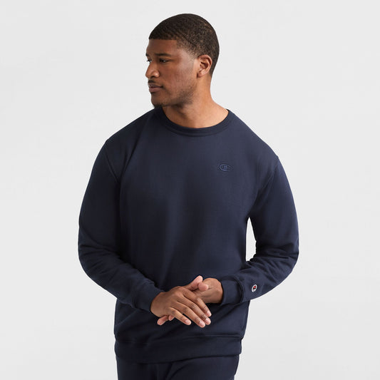 A man wears a navy Champion Powerblend Crewneck Sweatshirt, C Logo - Big & Tall, and matching pants, standing against a white background with his hands loosely clasped, looking to his right.