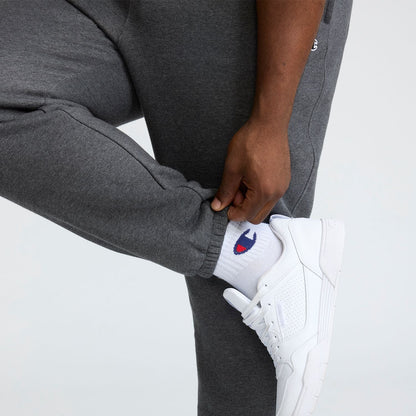 A person in Champion Powerblend Sweatpants, Cinched Bottom, C Logo - Big & Tall adjusts the cuff over white socks with a blue and red logo, paired with white sneakers against a plain light background.