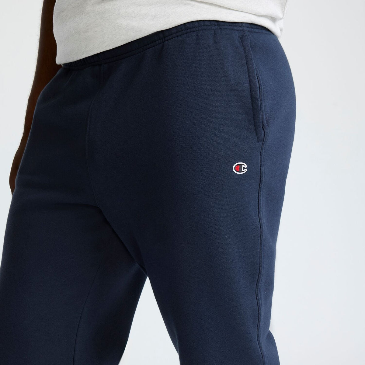 A person wearing navy blue Champion Powerblend Sweatpants, Open Hem, C Logo - Big & Tall with a light gray shirt is shown from the waist down, featuring the visible Champion logo on the pants.