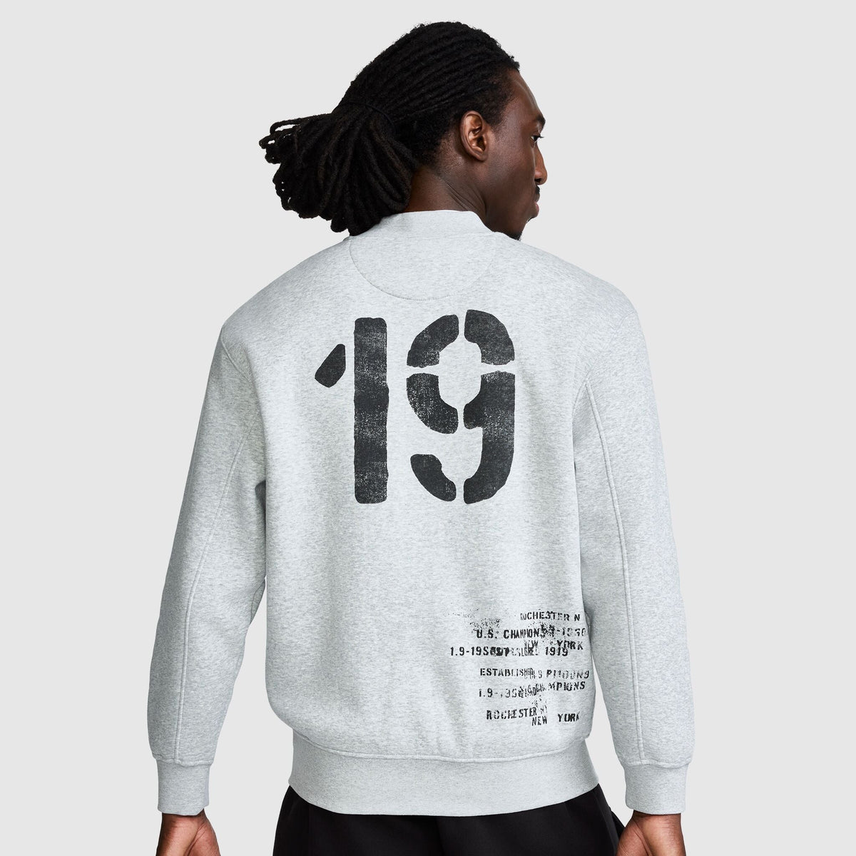 A person seen from the back wearing a Champion Fleece Baseball Jacket with the number 19 in large, distressed black print and smaller text near the bottom hem.