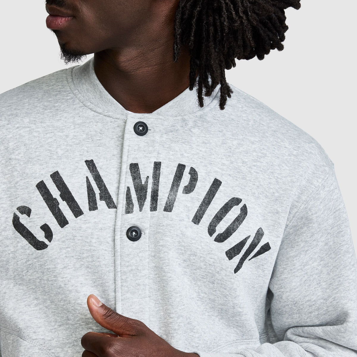Close-up of a person wearing a light gray snap-button Champion Fleece Baseball Jacket with the word CHAMPION printed in large, distressed black letters across the chest.