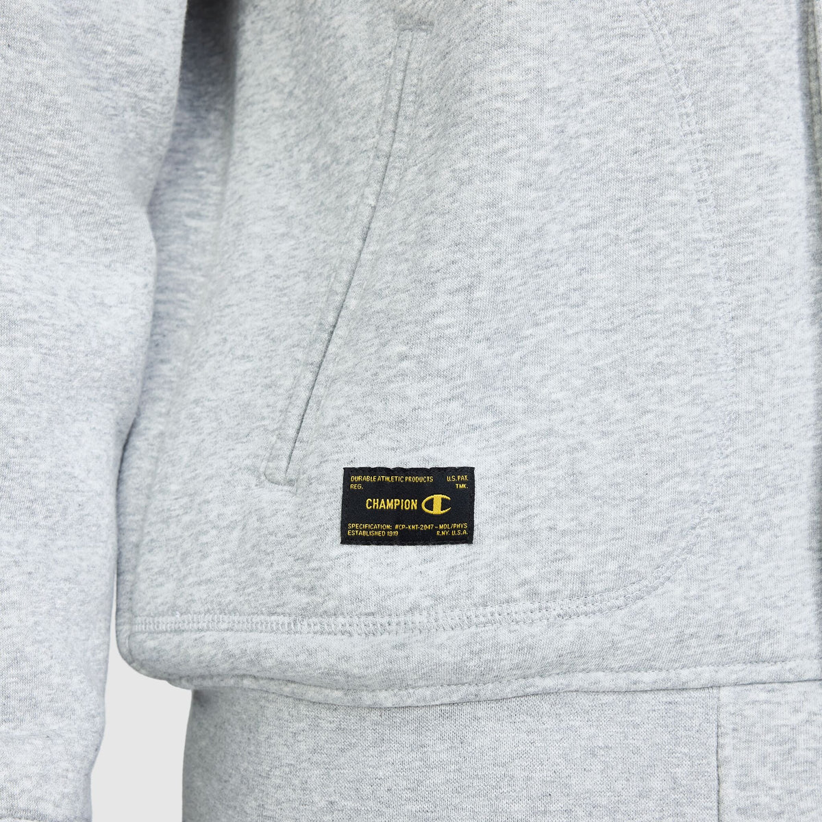 Close-up of a light gray Champion Fleece Baseball Jacket featuring a black rectangular patch with gold branding and technical text sewn onto the lower front pocket.