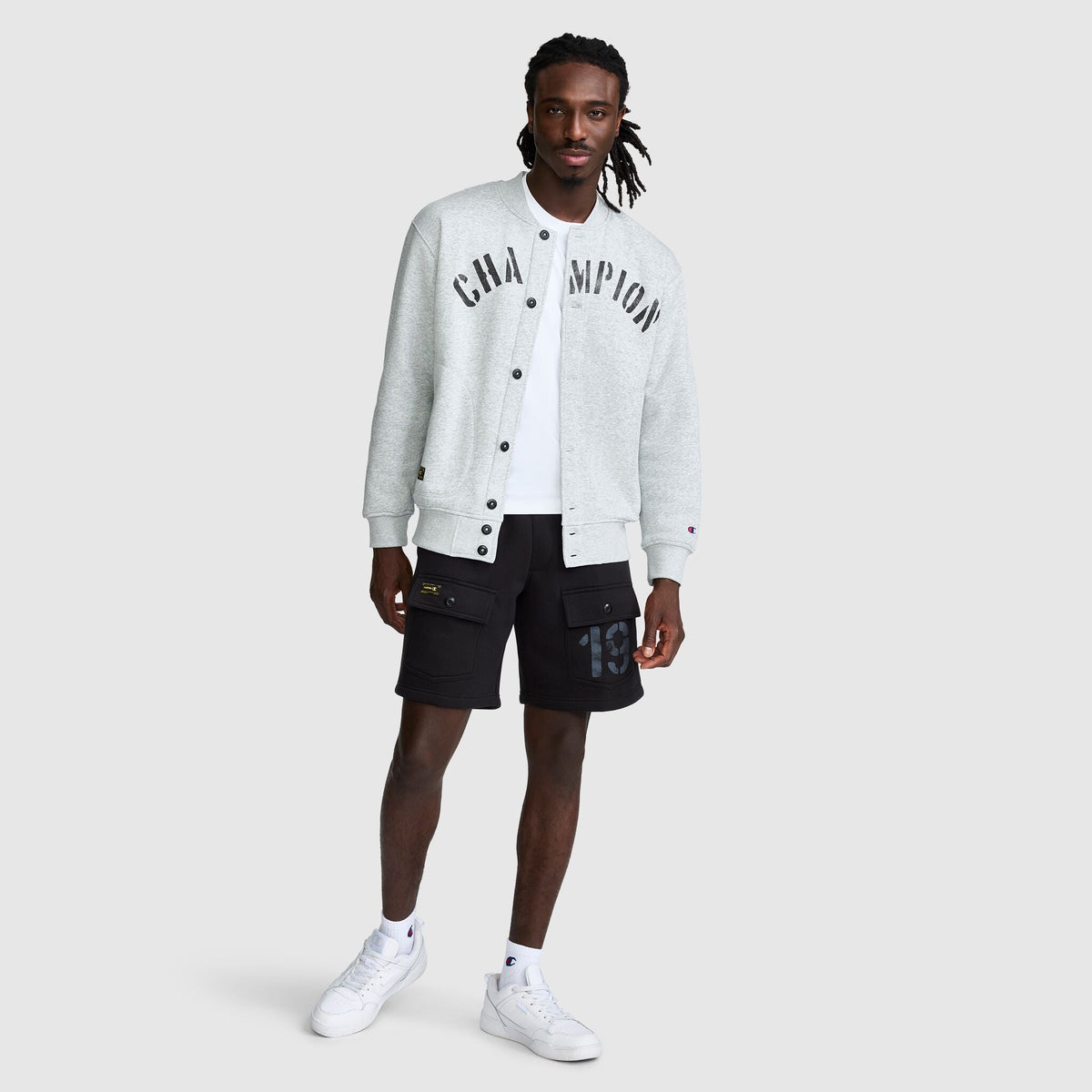 A man wears a grey Fleece Baseball Jacket with black lettering over a white t-shirt, paired with black cargo shorts and white sneakers.