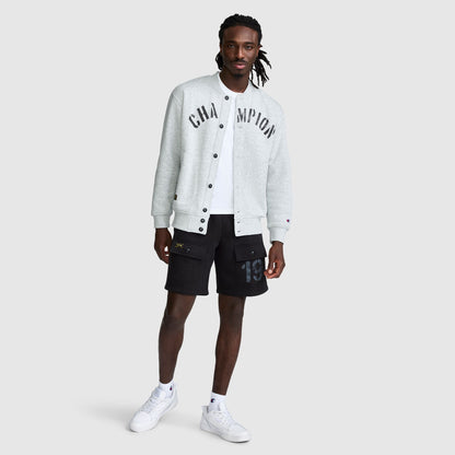 A man wears a grey Fleece Baseball Jacket with black lettering over a white t-shirt, paired with black cargo shorts and white sneakers.