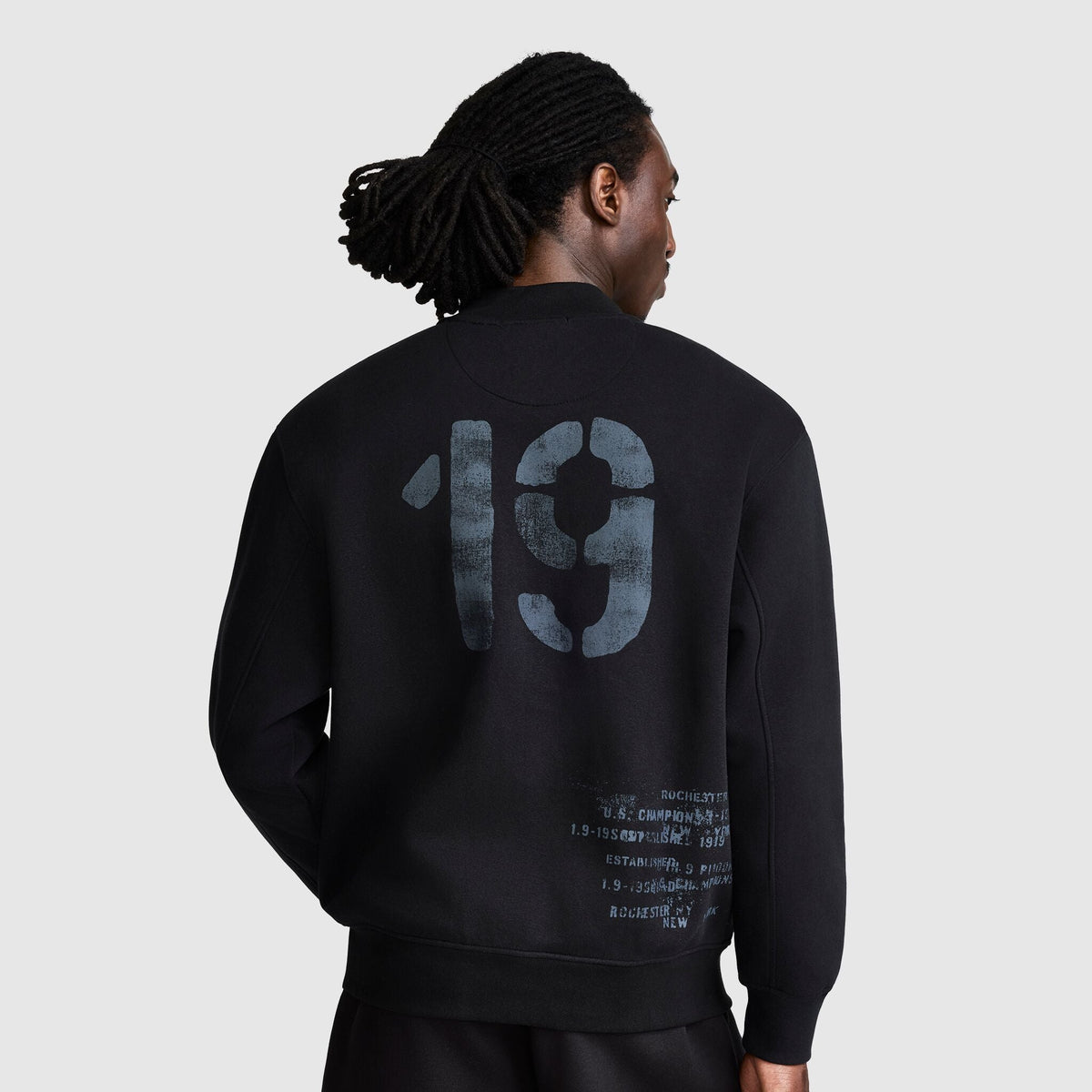 A person with long hair in a black Champion Fleece Baseball Jacket stands facing away, displaying a large faded 19 and smaller printed text on the back, evoking a vintage athletic style reminiscent of a classic varsity jacket.